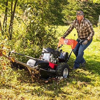 Man using self propelled brush hog clearing out bushes next to small tree